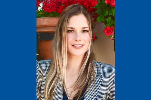 A photo of Veronica Russell, a young woman with long, straight blonde hair is smiling at the camera. She is wearing a houndstooth-patterned blazer and a silver necklace with a small pendant. The background features vibrant red flowers in a terracotta pot, adding a warm and natural setting to the image.